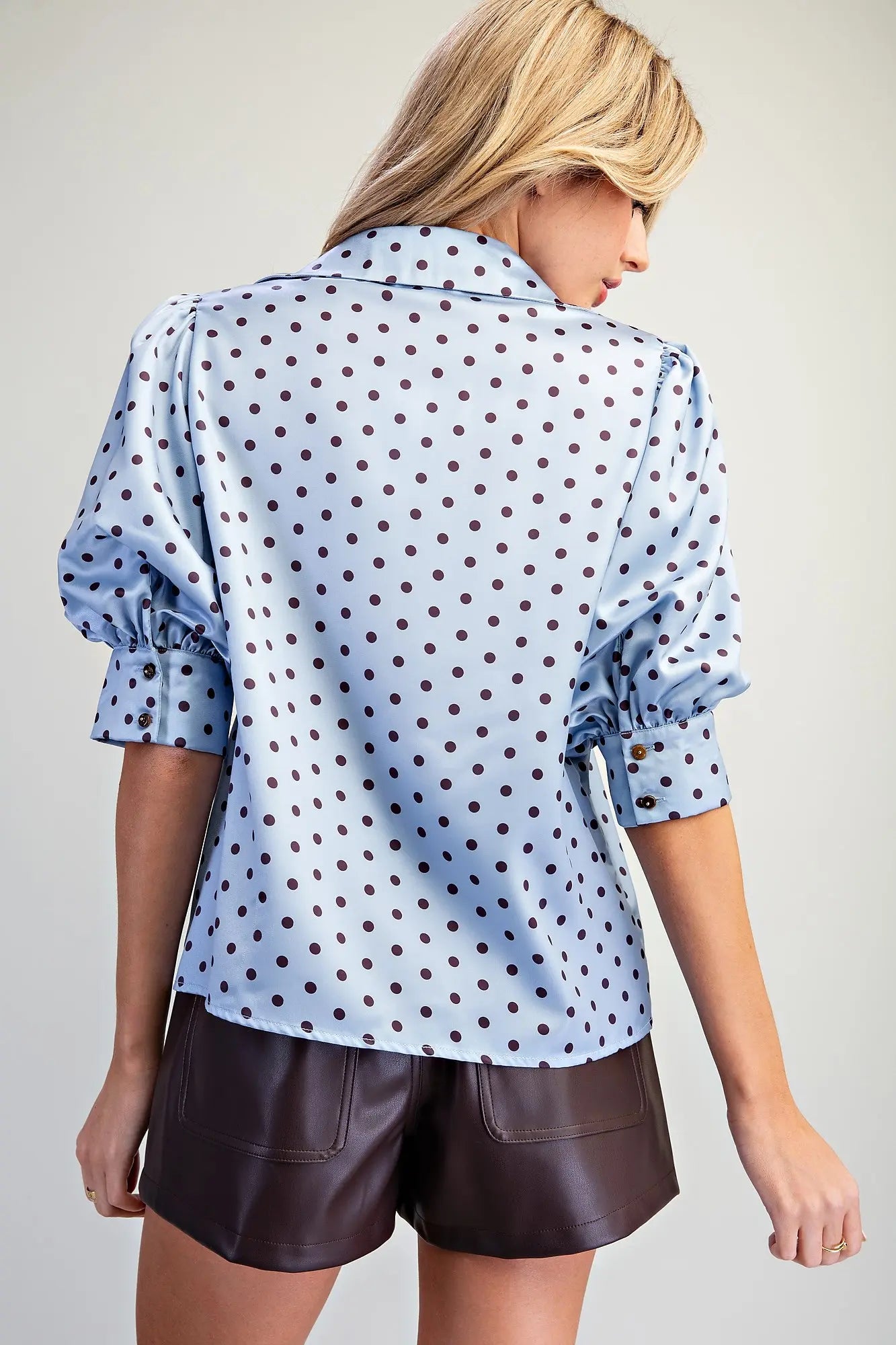 A woman shown from the back wearing a light blue satin blouse with dark brown polka dots and dark brown faux-leather shorts, set against a plain light gray studio background.