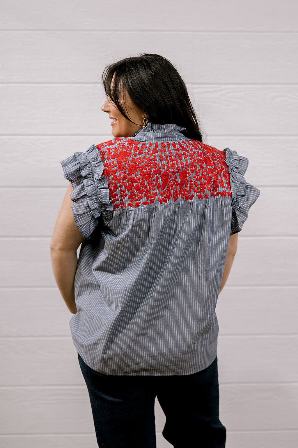 A woman shown from the back wearing a gray and white vertically striped blouse with ruffled sleeves and a red floral embroidered yoke, paired with dark pants, set against a white paneled wall.