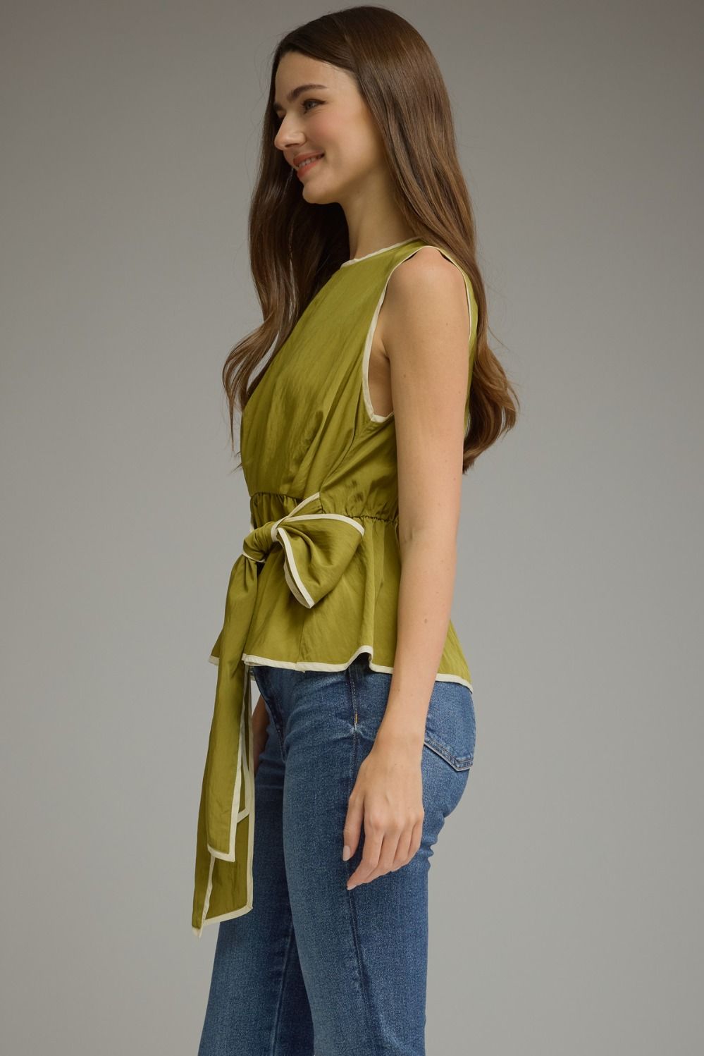 A woman wearing a sleeveless olive green top with a tie waist and blue jeans, shown from a side profile against a plain gray studio background.