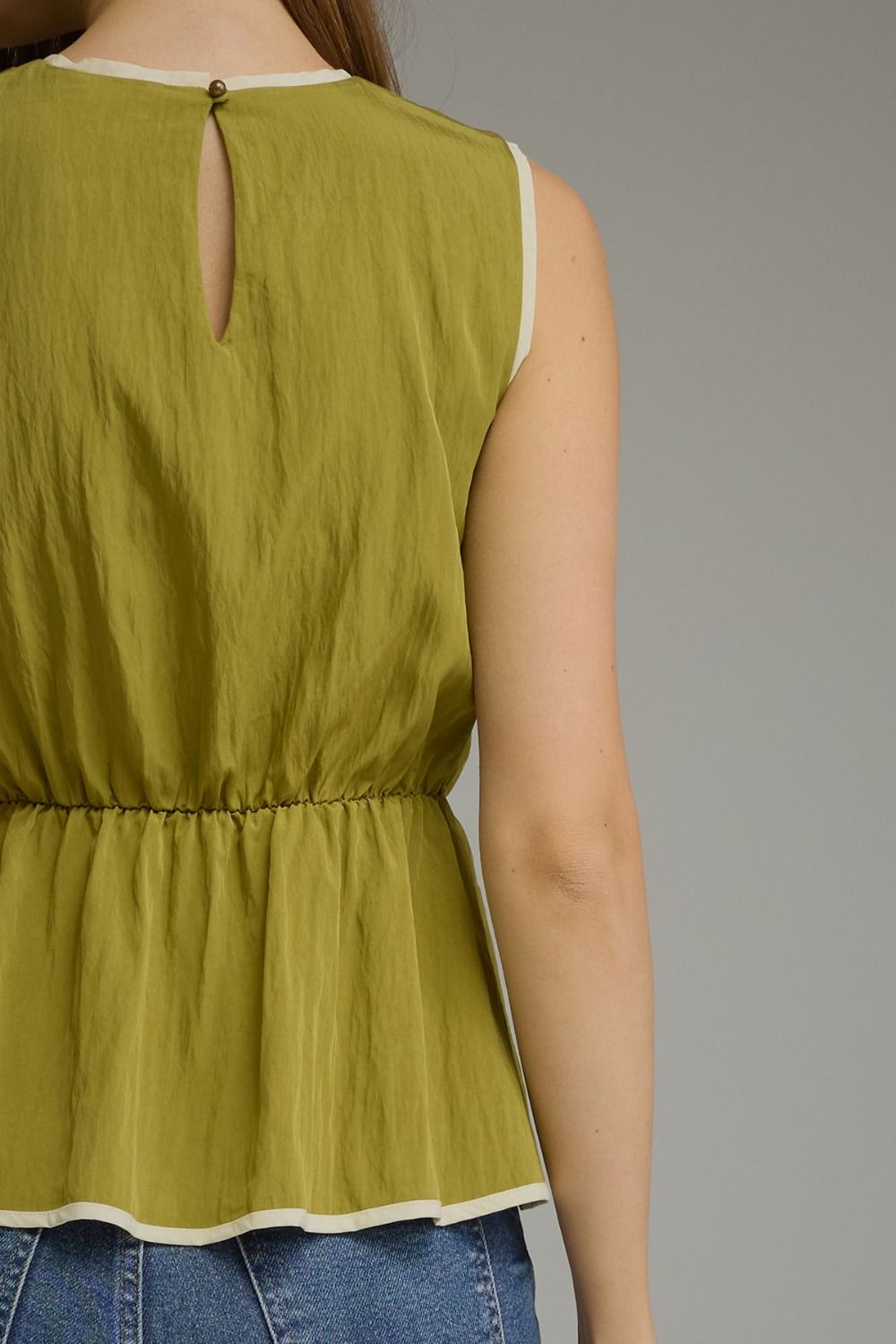 A woman wearing a sleeveless olive green peplum top with a keyhole back cutout and white trim, paired with blue jeans against a neutral gray background.