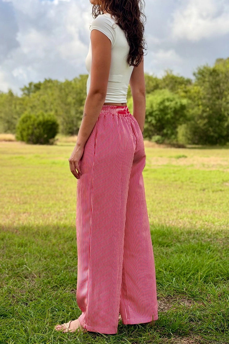 A woman standing barefoot in a grassy field, wearing high-waisted, wide-leg pink pleated pants and a white sleeveless top.