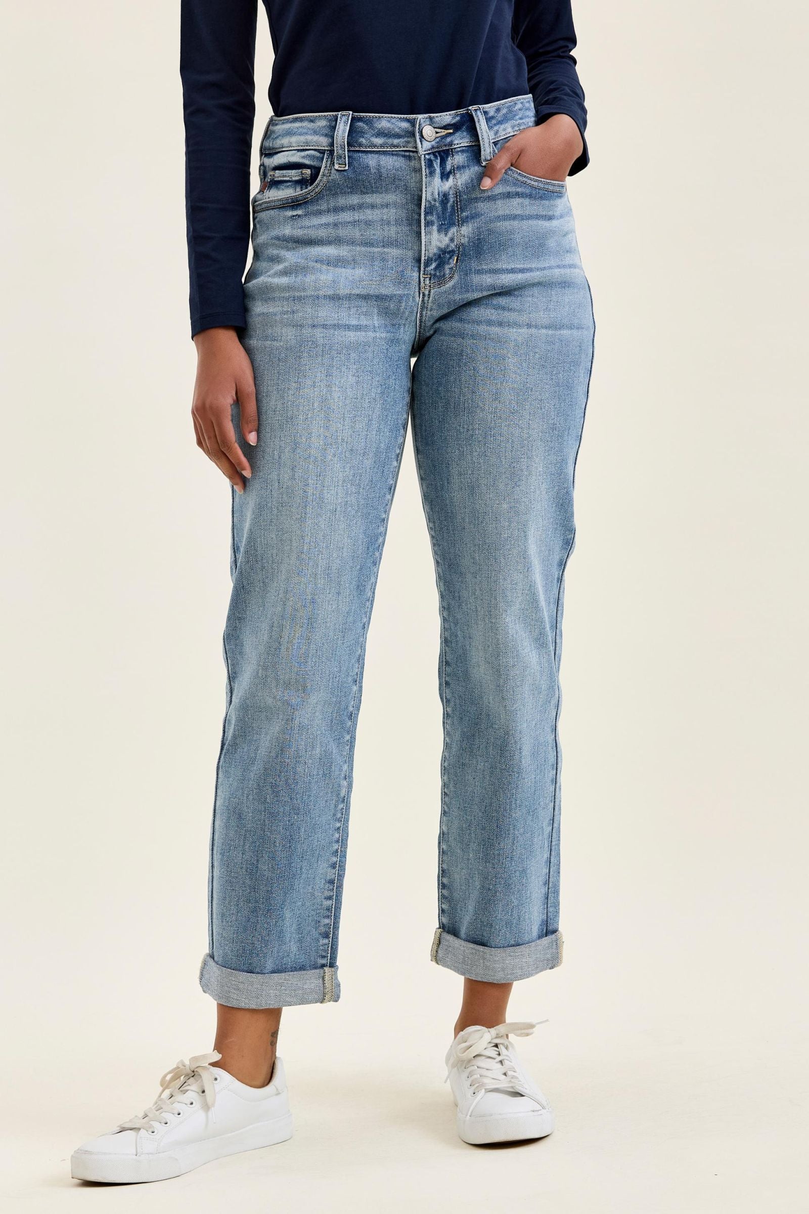 A person wearing light-wash blue denim jeans with a straight-leg fit, cuffed at the ankles, paired with a navy long-sleeve top and white sneakers against a plain light beige studio background.