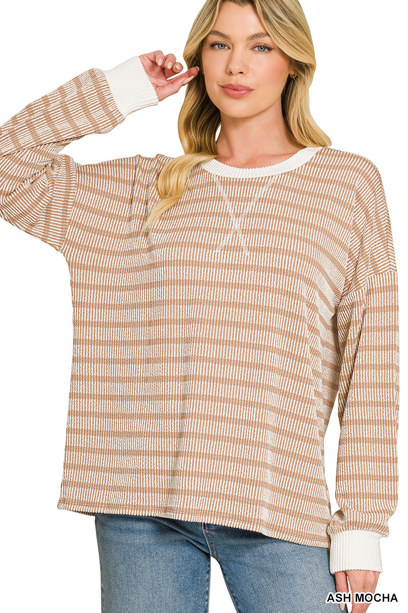 A woman modeling a long-sleeve knit top in a light ash mocha color with fine horizontal stripes and contrasting white ribbed cuffs and collar, paired with blue jeans against a white background.