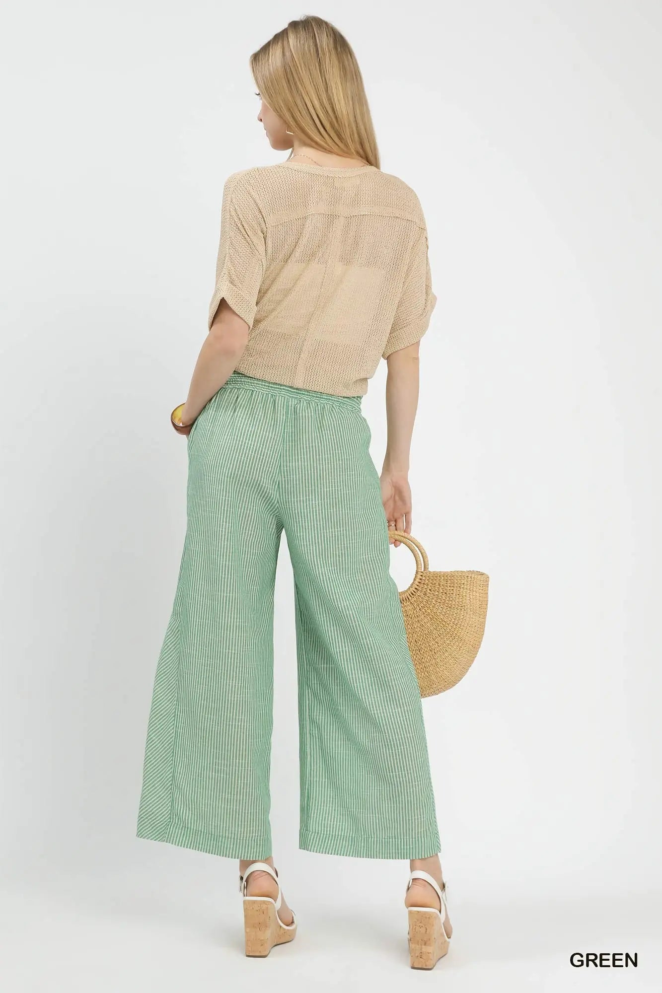 Woman wearing a beige top and green checkered pants, holding a straw bag on a white background.