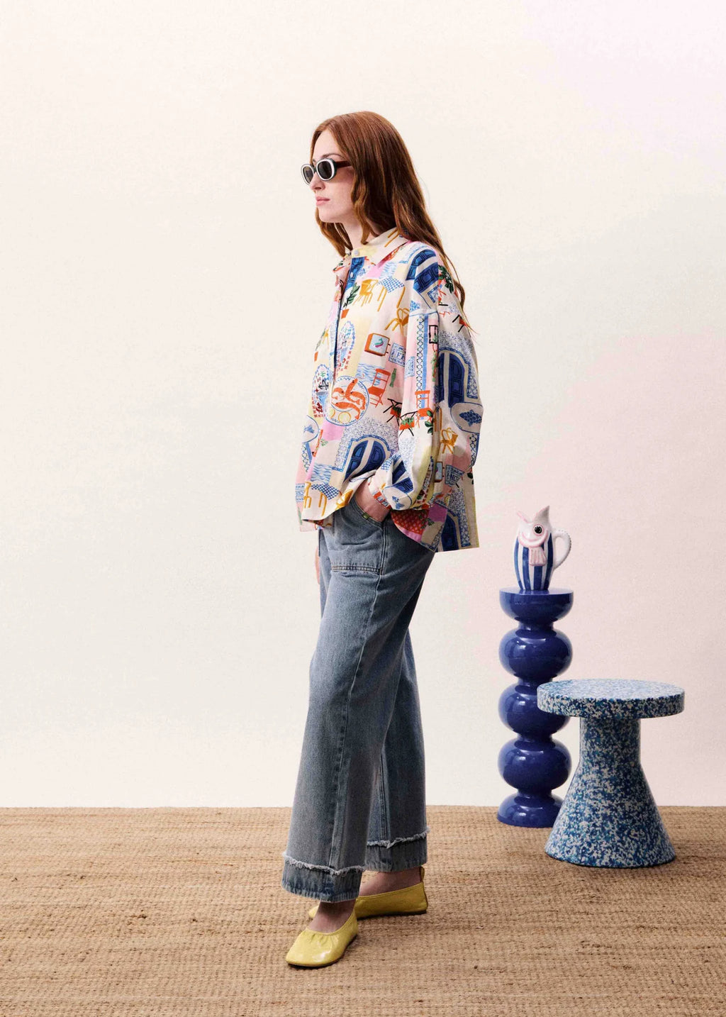 Person wearing a colorful jacket and jeans standing in a room with a white wall and two small tables.