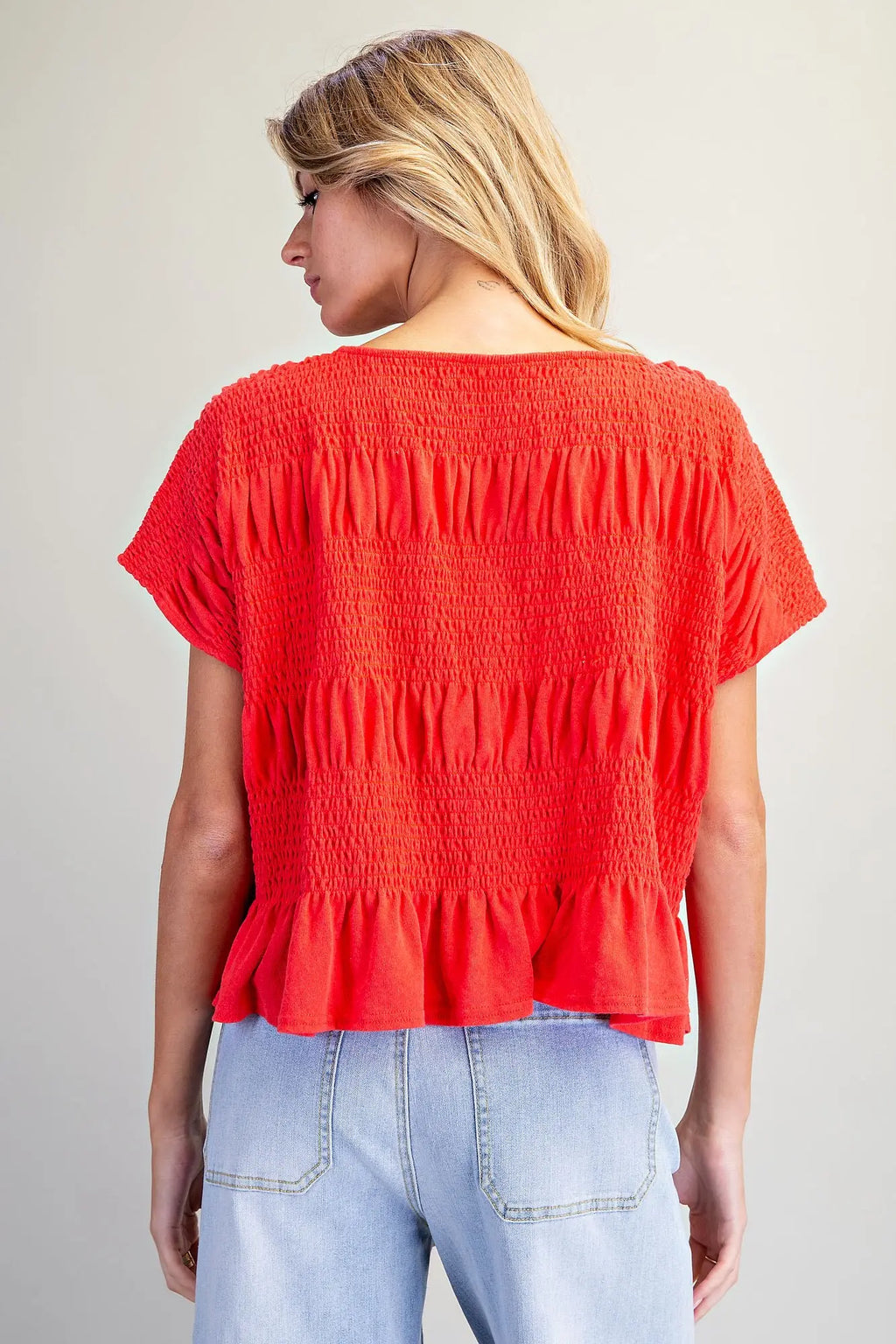 A woman shown from the back wearing a bright red short-sleeve smocked blouse with a ruffled hem, paired with light blue jeans, against a plain light gray background.