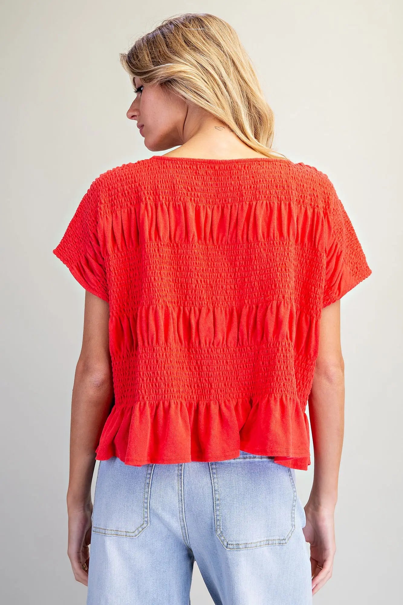 A woman shown from the back wearing a bright red short-sleeve smocked blouse with a ruffled hem, paired with light blue jeans, against a plain light gray background.