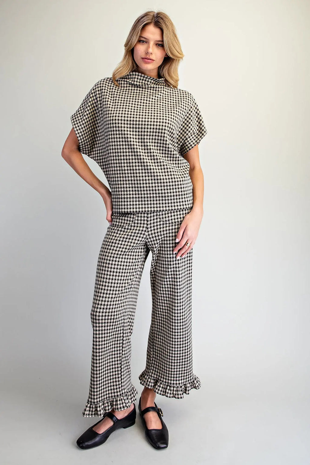 A woman modeling a matching two-piece outfit featuring a black and white gingham patterned short-sleeve mock-neck top and matching wide-leg trousers with ruffled hems, paired with black Mary Jane shoes, against a plain light gray studio background.