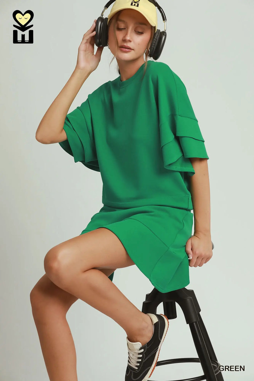 Person wearing a green outfit with headphones and a cap, sitting on a stool against a plain background.