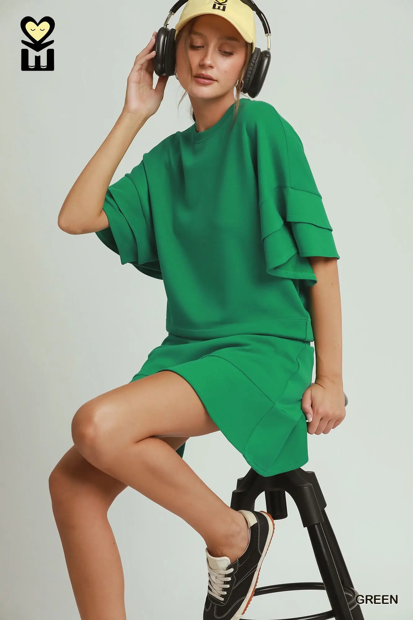 Person wearing a green outfit with headphones and a cap, sitting on a stool against a plain background.