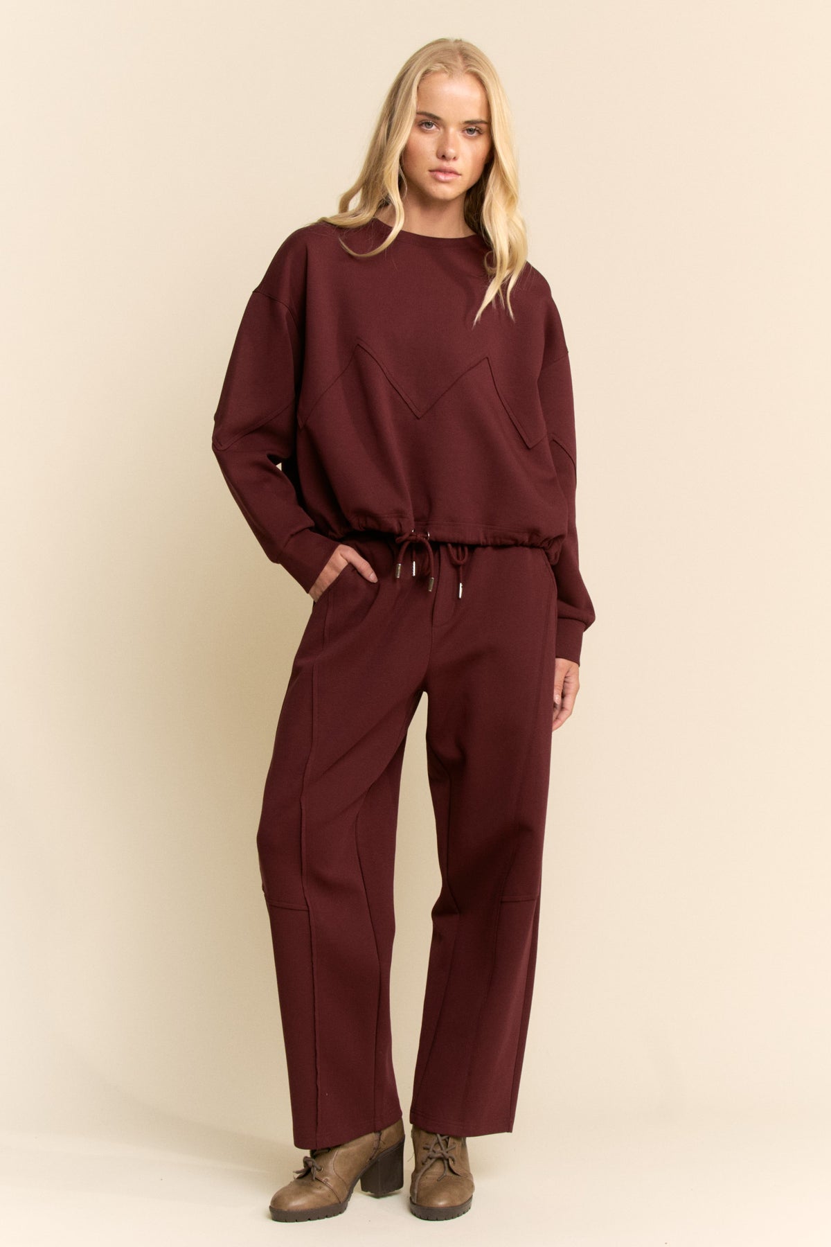 A blonde model wearing a matching burgundy sweatshirt and wide-leg sweatpants set with brown shoes, standing against a solid beige studio background.