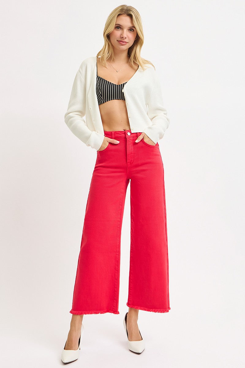 A woman modeling bright red wide-leg cropped jeans with a white cropped cardigan, a black and white striped crop top, and white pointed-toe heels against a plain white studio background.