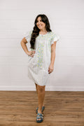 Woman wearing a white dress with floral patterns standing against a white brick wall.