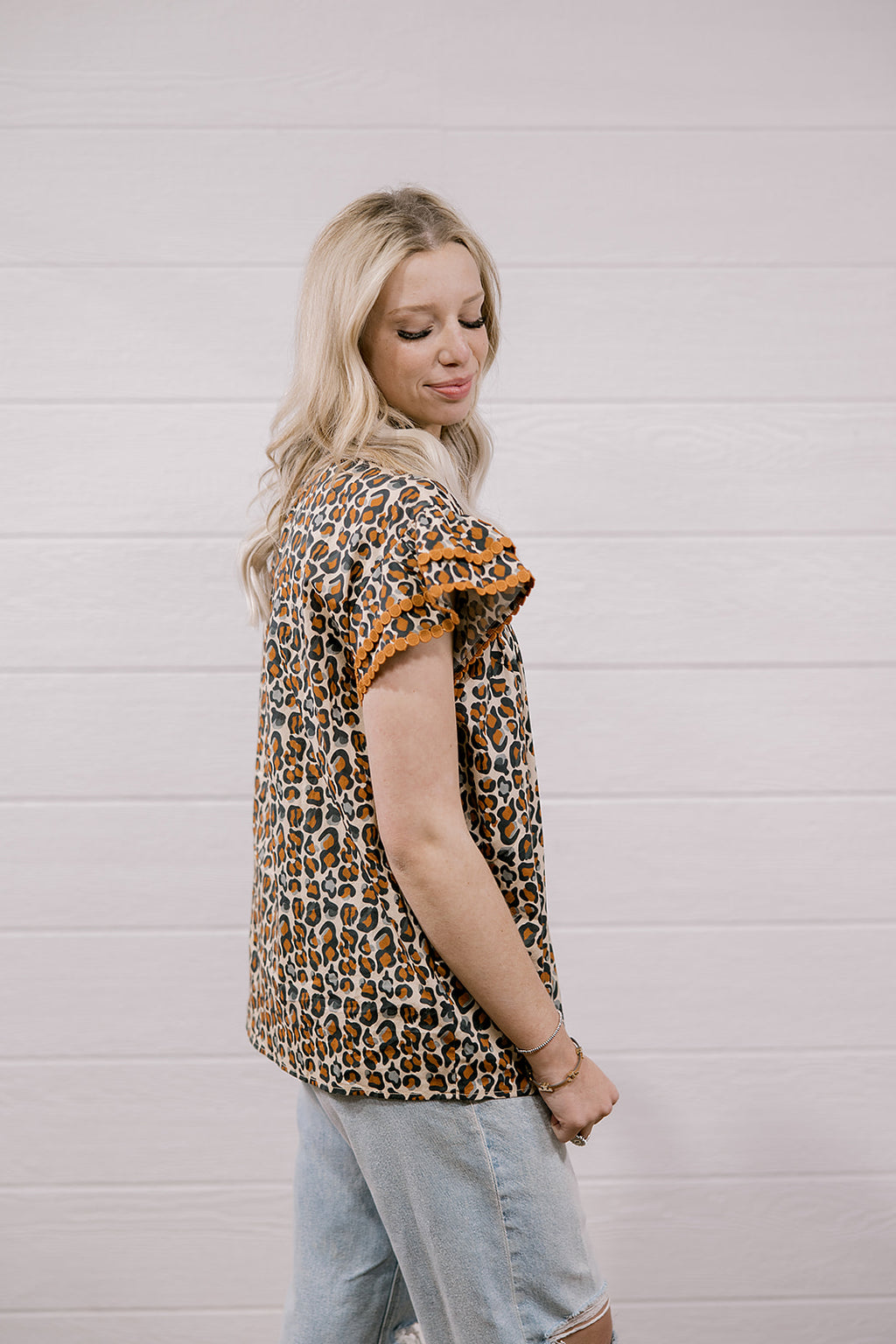 A woman wearing a leopard print short-sleeve top with ruffled sleeves and light-wash denim shorts, standing against a white wood-paneled wall.