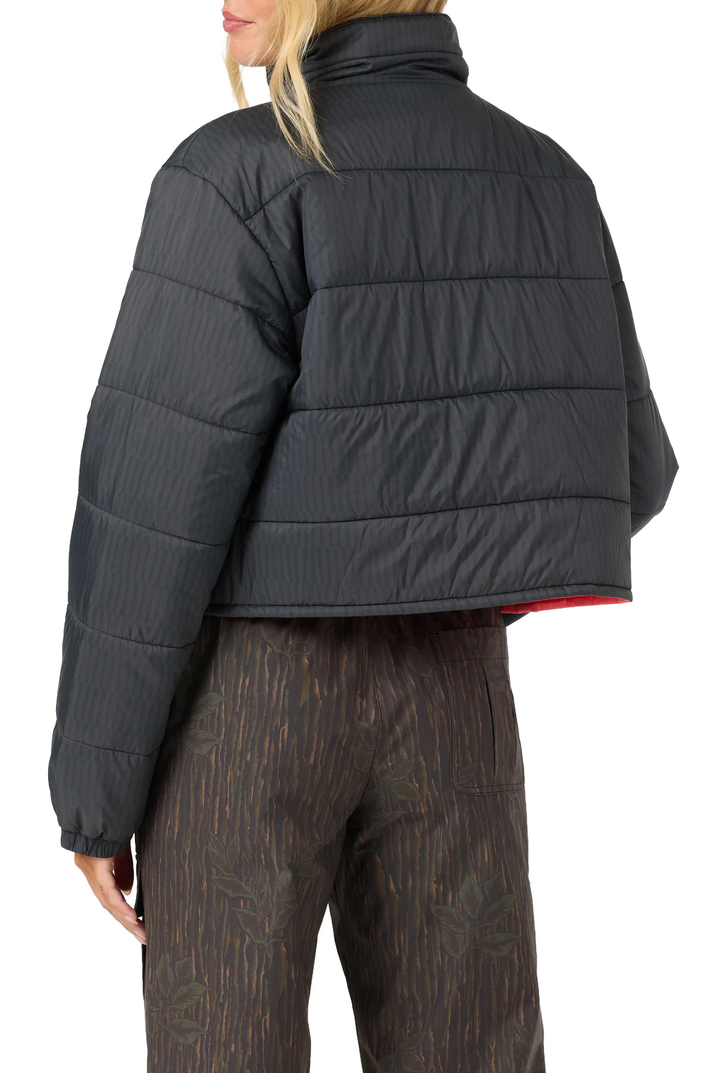 A woman shown from the back wearing a cropped black quilted puffer jacket with a subtle vertical texture and a red lining visible at the hem, paired with brown patterned pants against a white background.