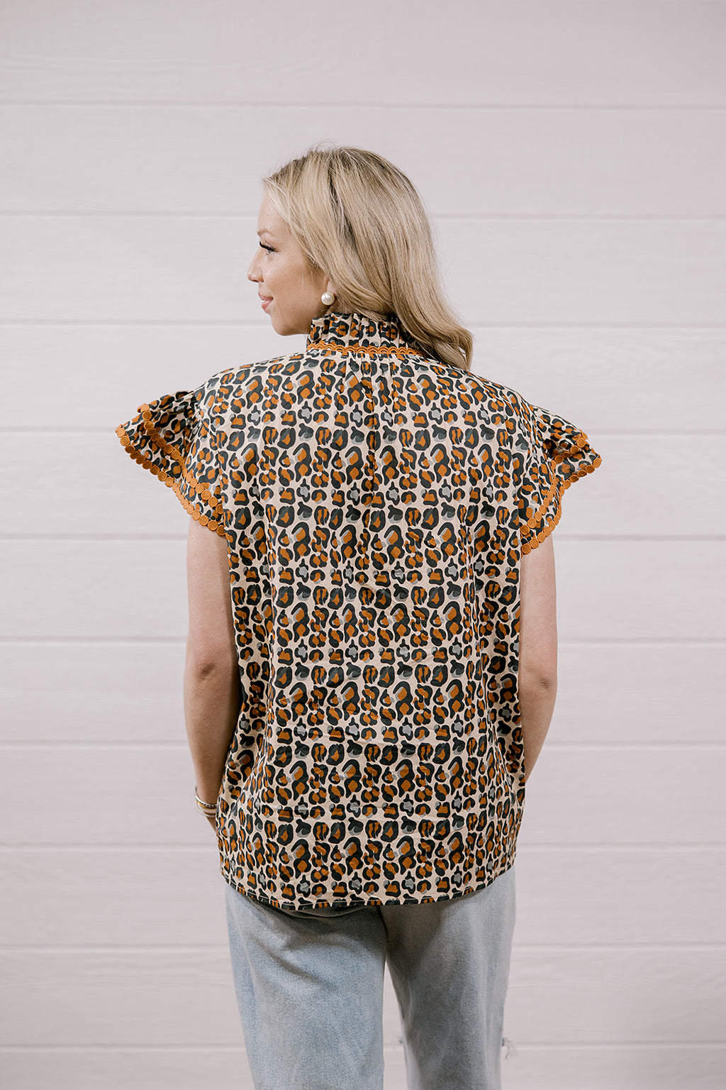 A woman shown from the back wearing a leopard print short-sleeve blouse with ruffled sleeves and light blue jeans, standing against a white paneled wall.