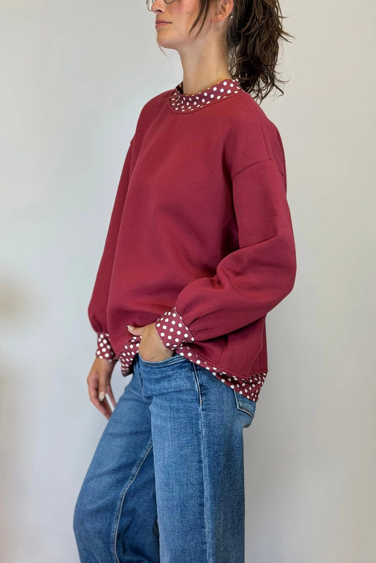 A woman wearing a burgundy crewneck sweatshirt with red and white polka dot trim on the collar and cuffs, paired with blue jeans, shown from a side profile against a plain light background.