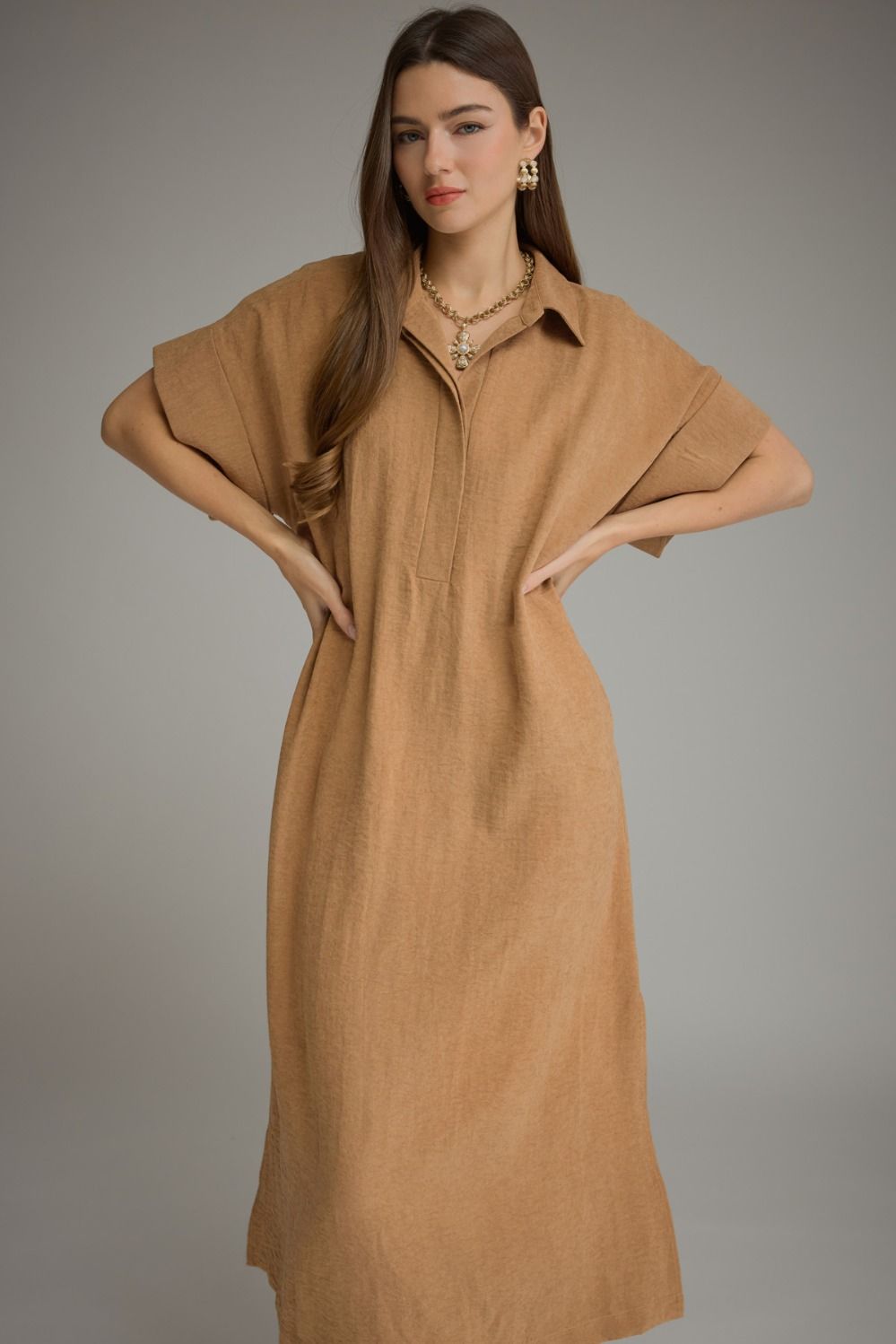 A woman modeling a loose, tan-brown linen midi dress with a collar and short wide sleeves, posed against a neutral gray studio background.