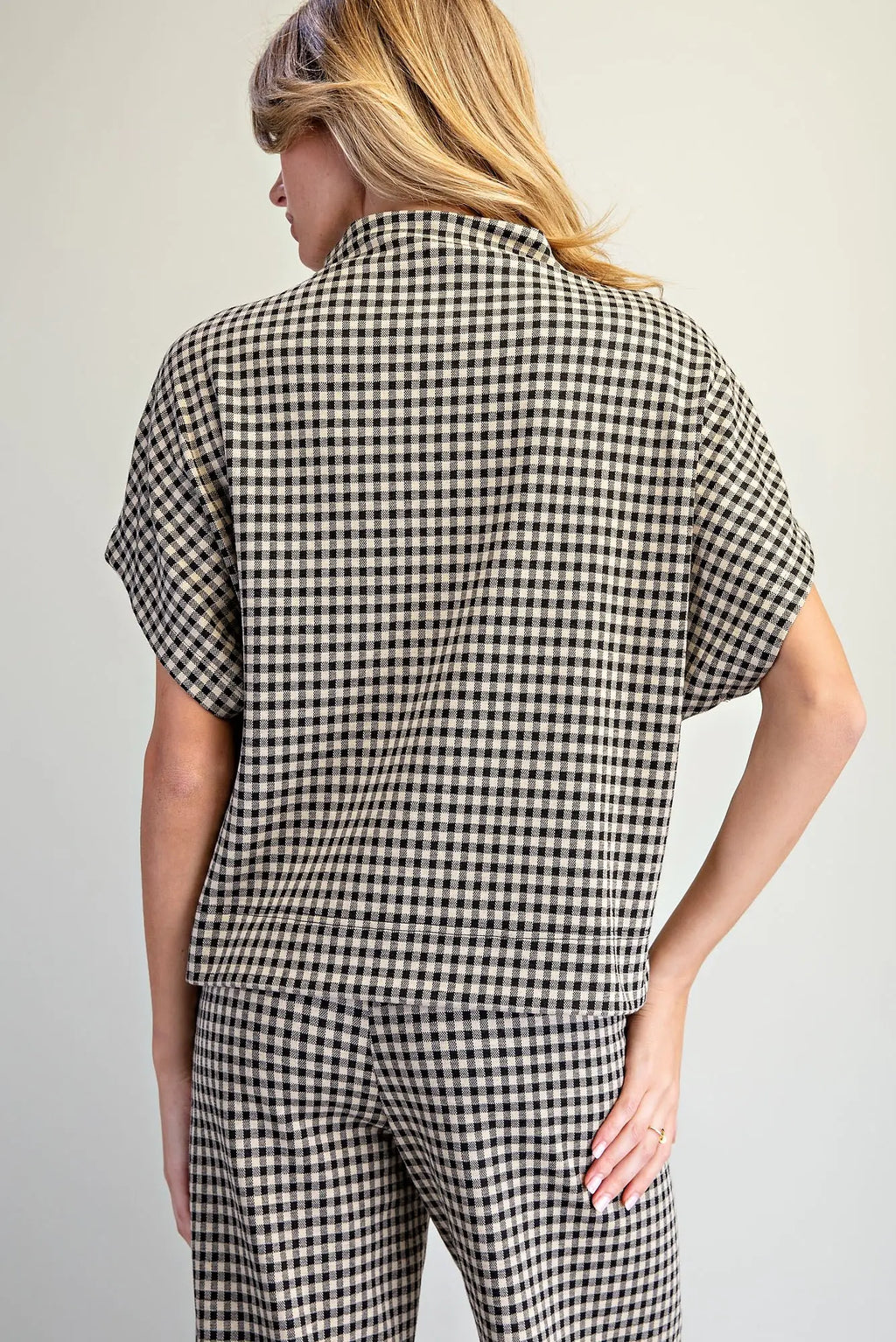 A woman shown from the back wearing a short-sleeve collared shirt and matching pants in a black and beige gingham check pattern, against a plain light gray studio background.