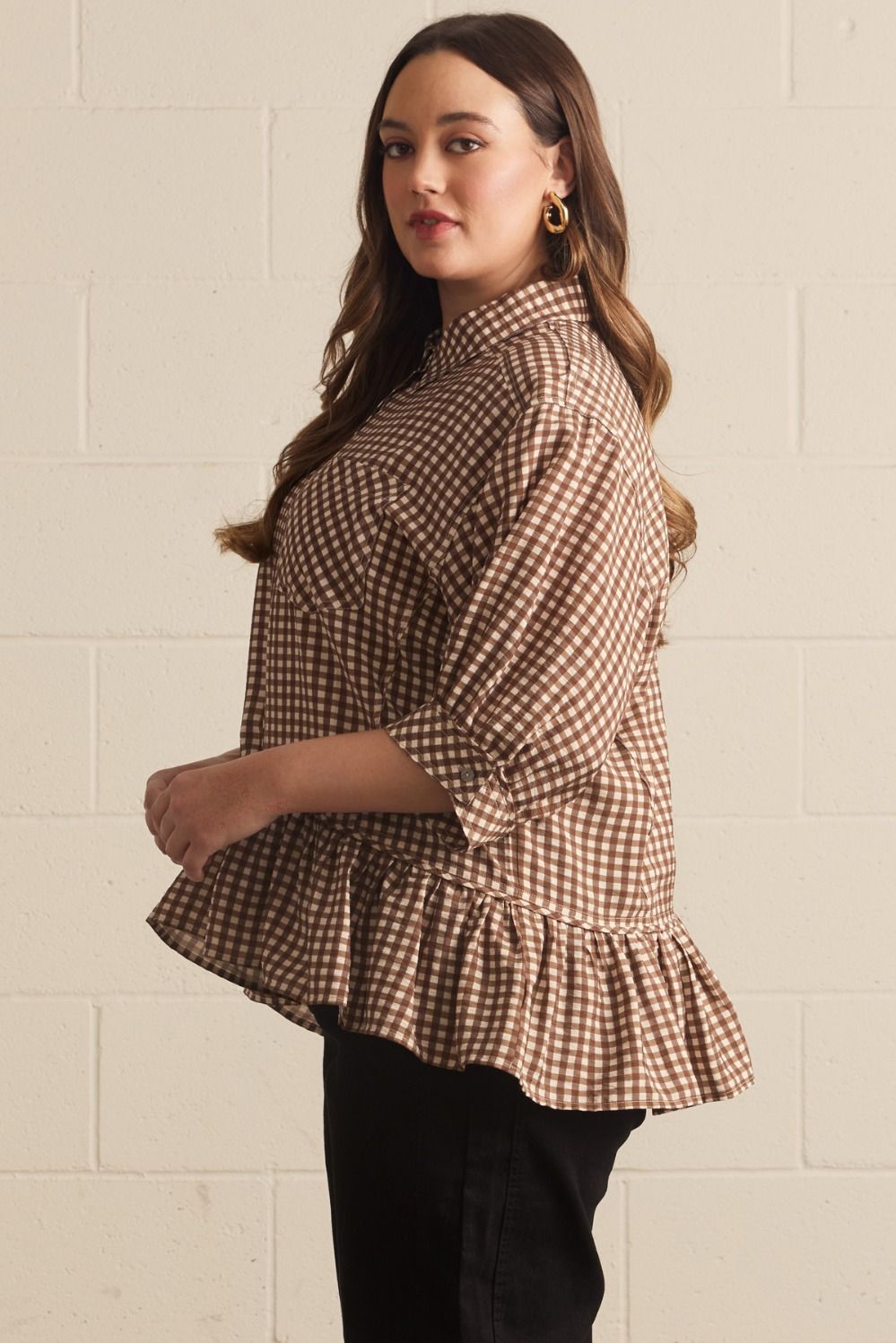 Woman wearing a brown checkered blouse with ruffled sleeves against a beige brick wall.