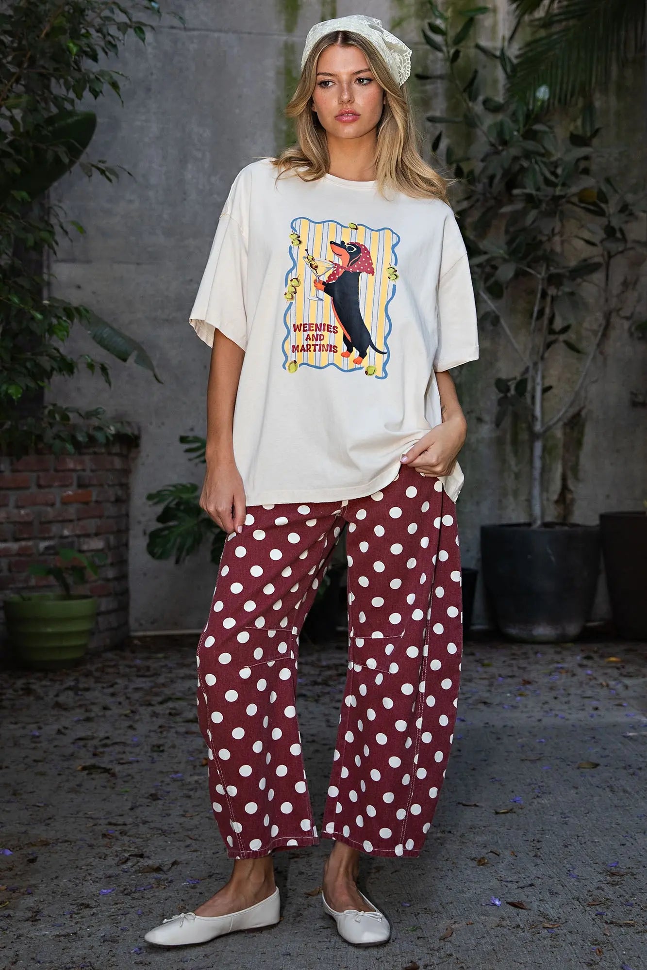 A woman stands outdoors wearing an oversized cream graphic T-shirt with a colorful circus-themed print, burgundy wide-leg pants with white polka dots, and white flats. She is posed in front of a gray wall with potted plants.