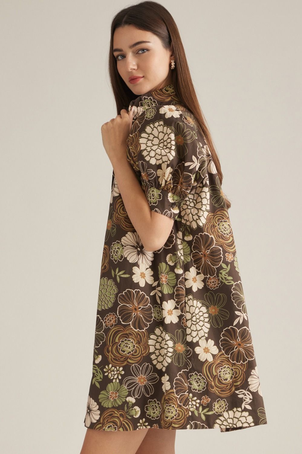 A woman modeling a short-sleeve high-neck dress with a brown, beige, and green floral pattern, photographed against a solid light gray background.