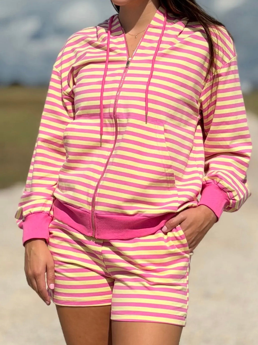A woman wearing a pink and yellow striped zip-up hoodie and matching shorts, photographed outdoors with a blurred natural background.