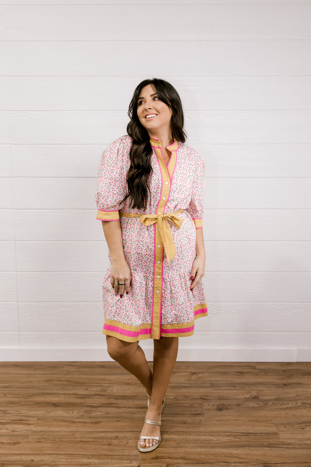 Woman wearing a patterned dress with a yellow belt against a white wooden panel background