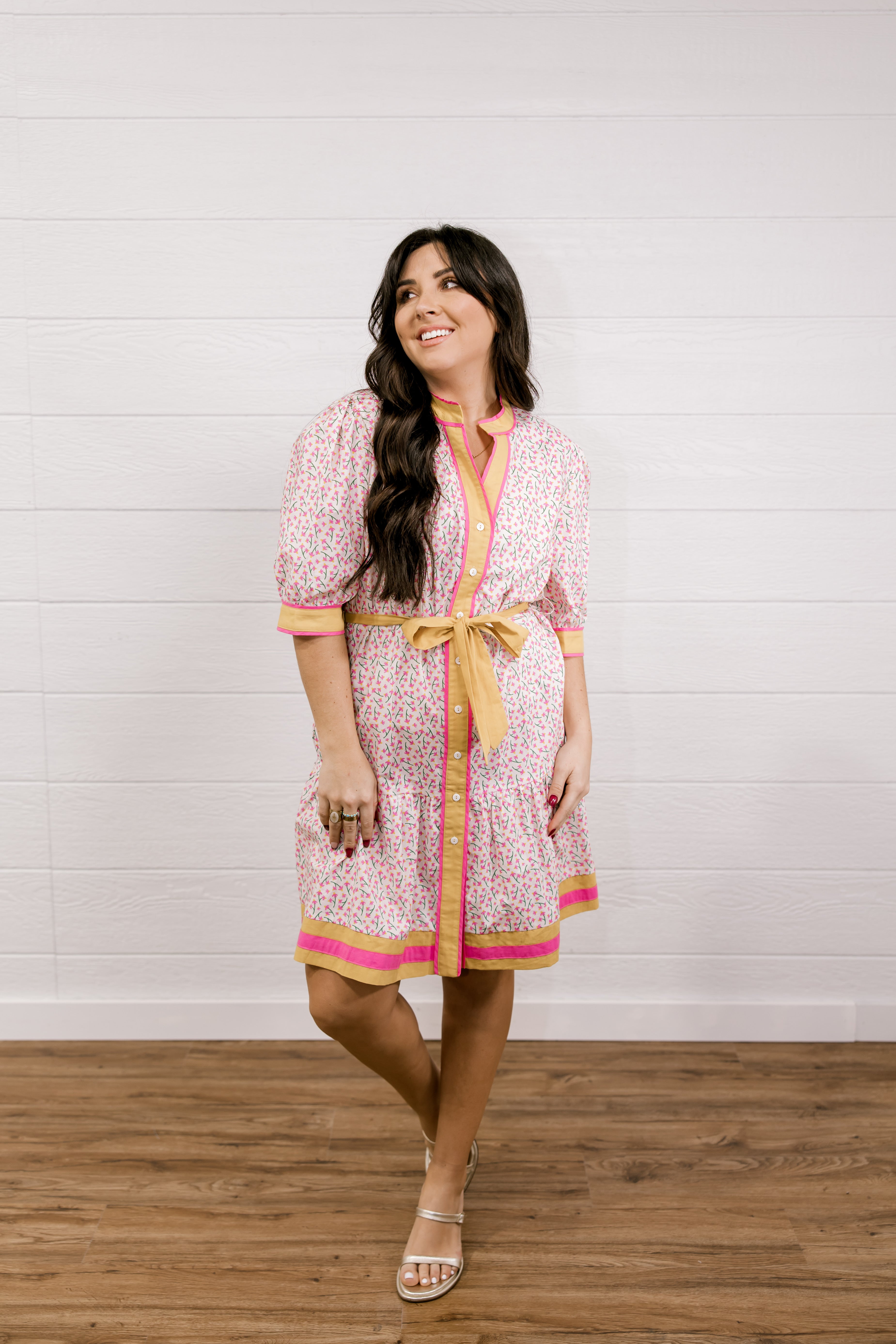 Woman wearing a patterned dress with a yellow belt against a white wooden panel background