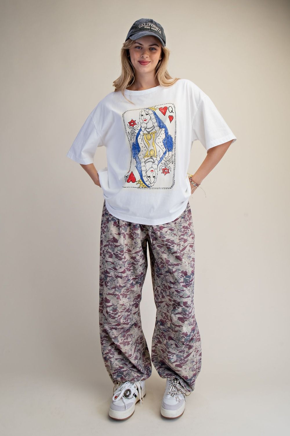 Person wearing a white t-shirt with a graphic design, patterned pants, and a cap on a plain background