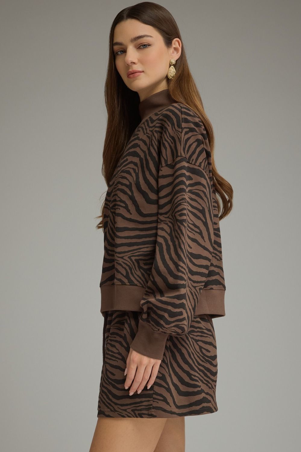 A woman modeling a brown and black zebra-print mock-neck long-sleeve sweatshirt and matching mini skirt set against a solid gray studio background.