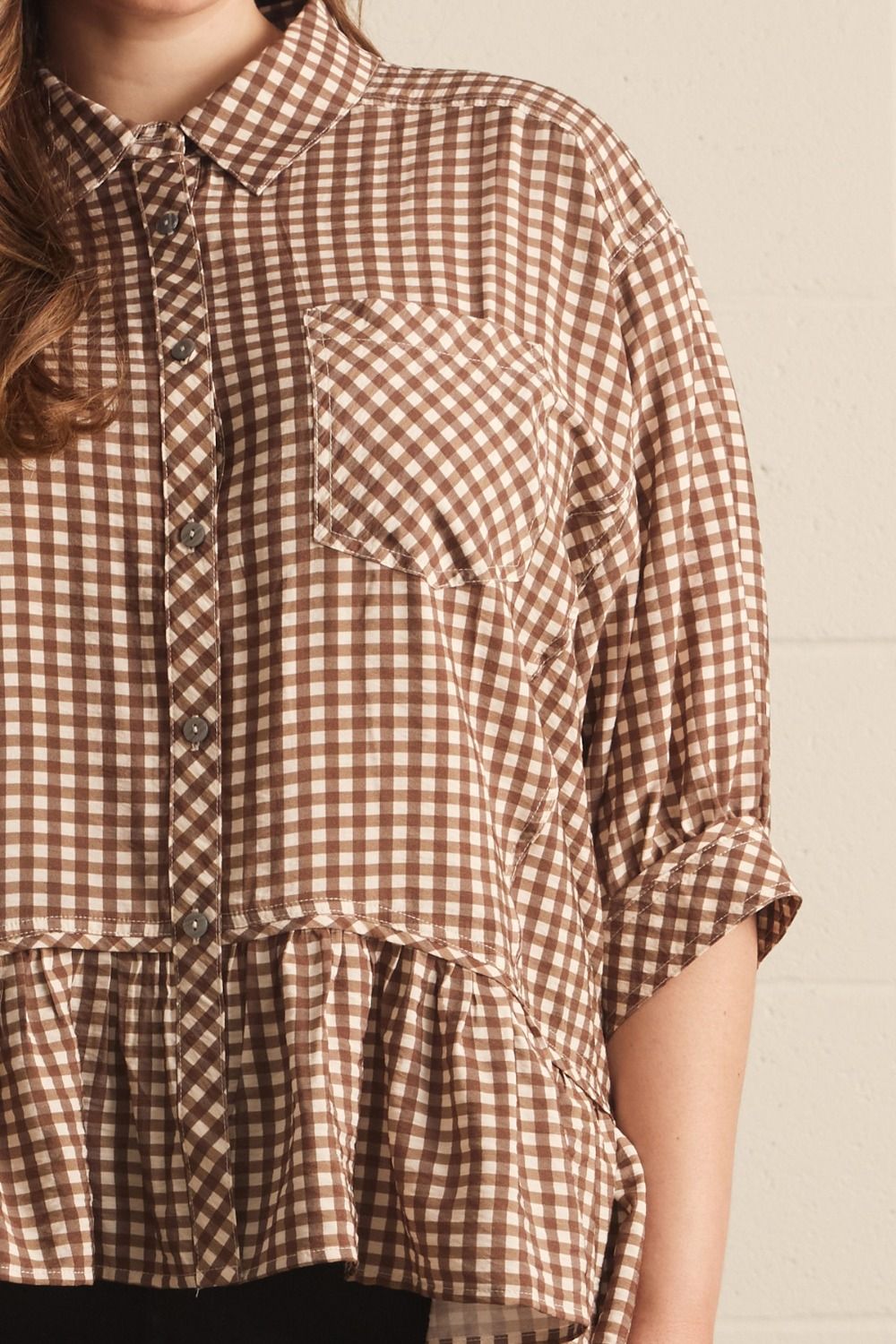 A close-up of a woman wearing a brown and beige gingham check button-up shirt with a ruffled hem and three-quarter sleeves.