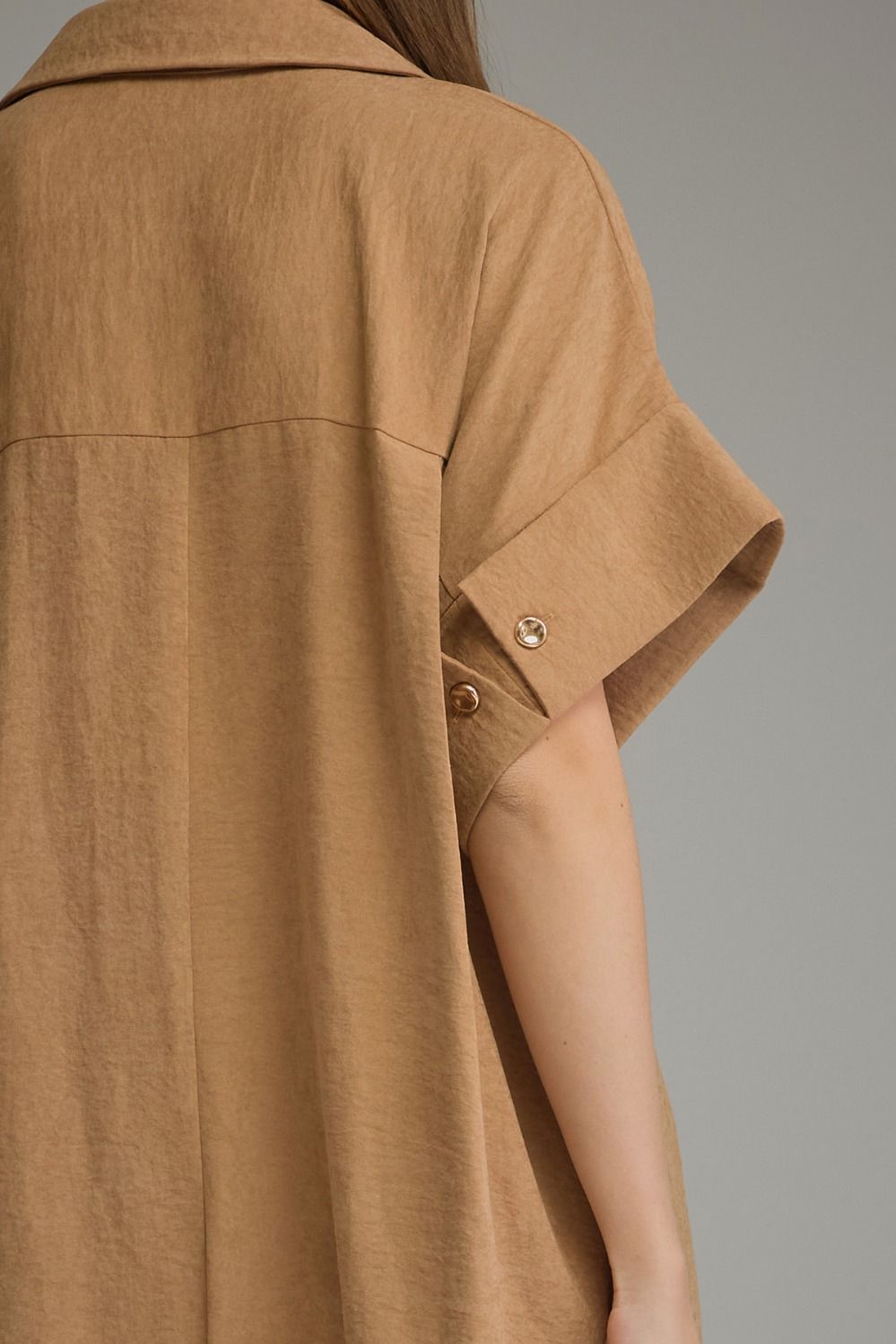 Close-up of a person wearing a loose tan short-sleeve shirt or tunic with rolled cuffs and visible buttons, shown against a plain gray studio background.