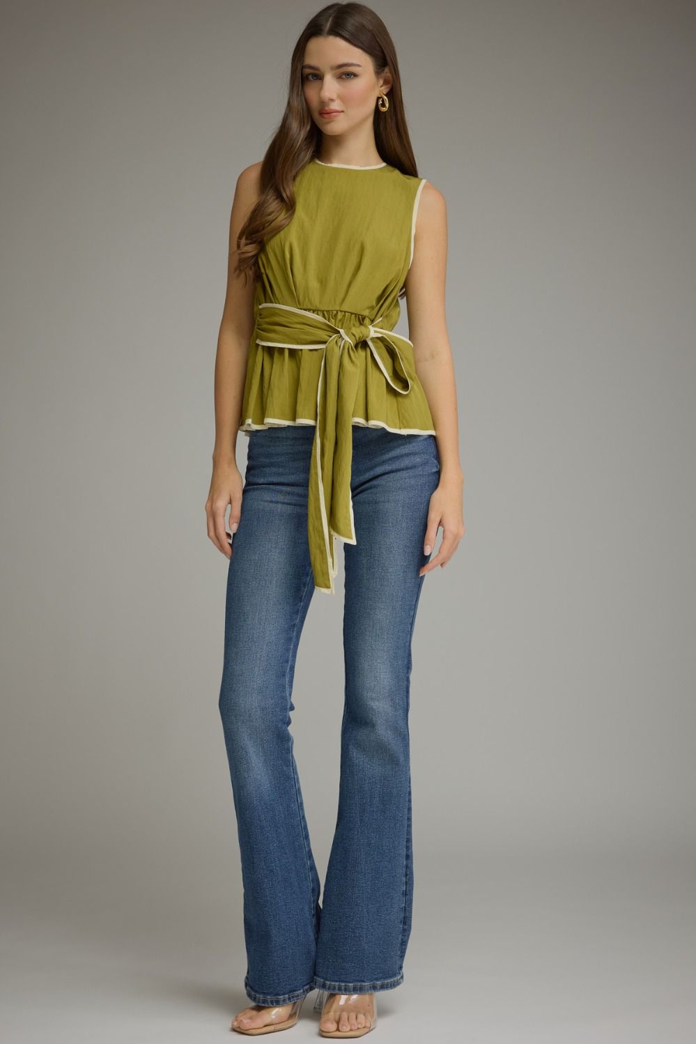 A woman modeling a sleeveless olive green peplum top with a matching tie belt and blue flare jeans against a neutral gray studio background.