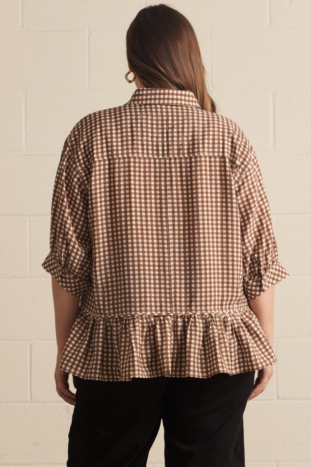 Person wearing a brown checkered blouse with ruffled details on a beige brick wall background