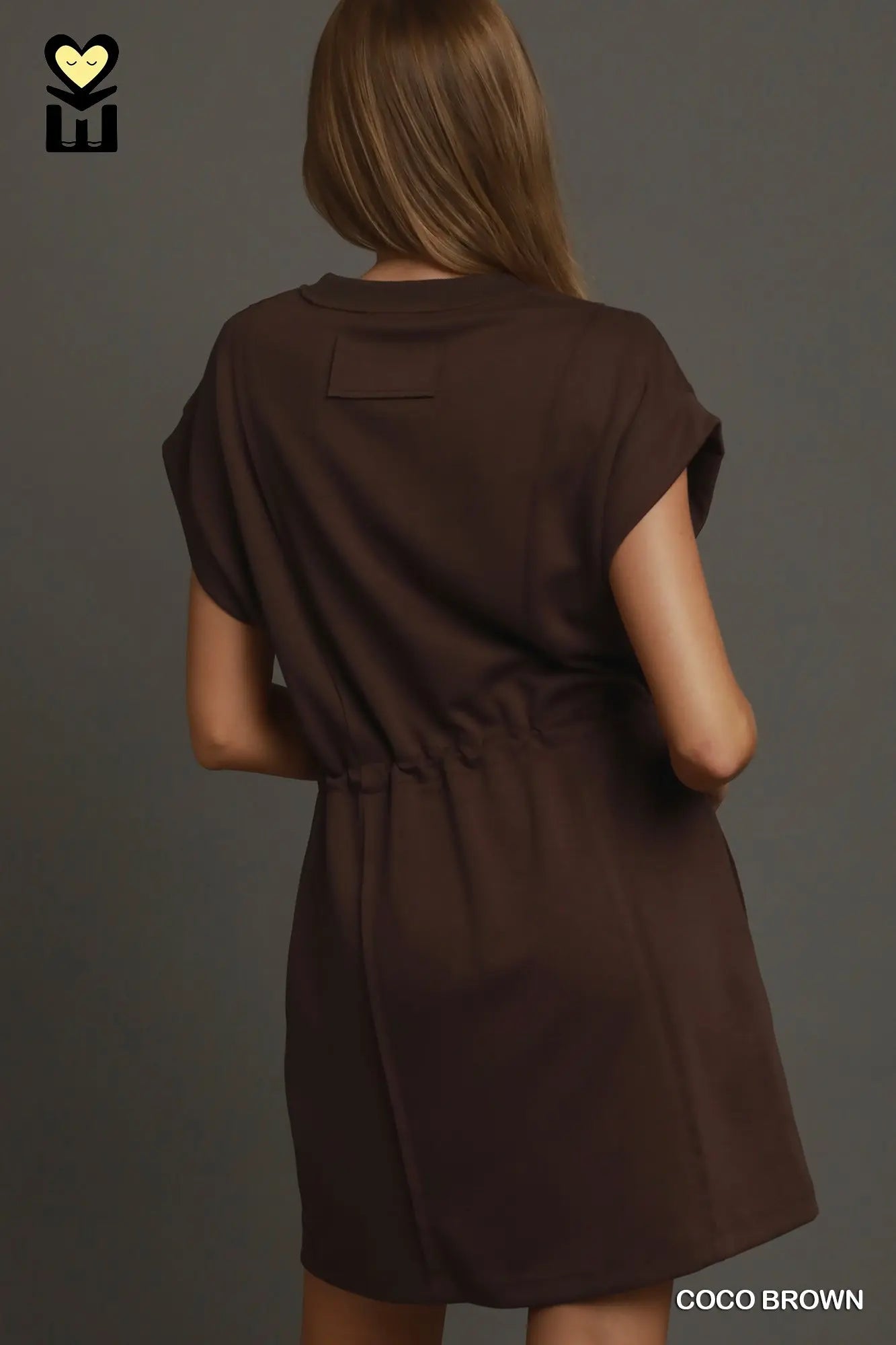 A woman shown from the back wearing a short-sleeve dark brown dress with a gathered waist, photographed against a dark gray studio background. The text 'COCO BROWN' appears in the bottom right corner.