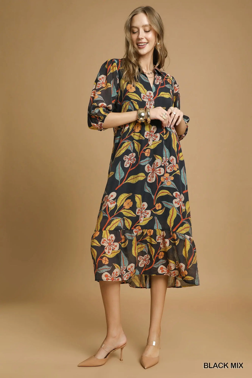 A woman modeling a black midi dress with a colorful floral print, standing against a solid tan studio background. She is wearing beige high heels and gold bracelets.