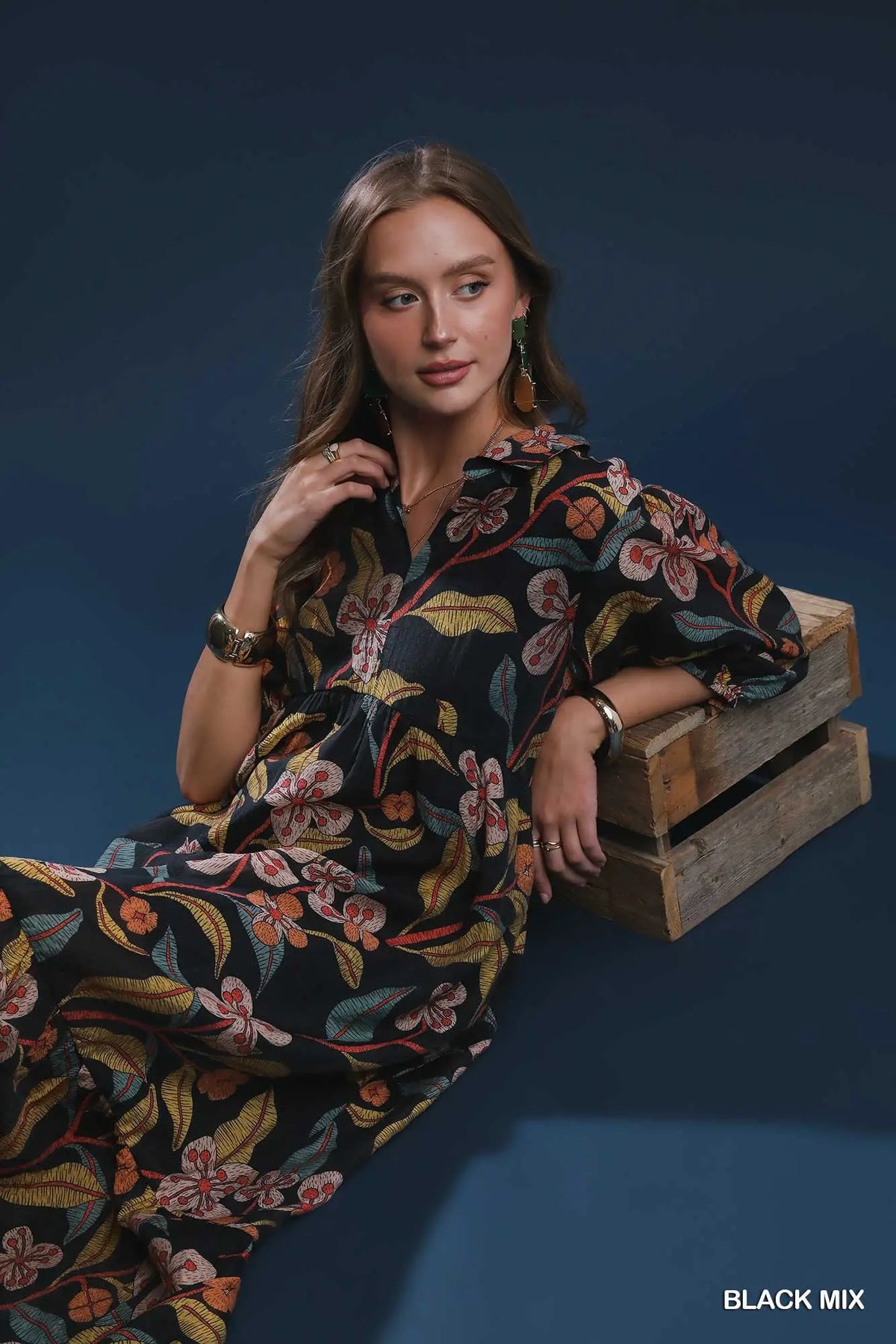 A woman models a black floral dress while leaning on stacked wooden crates against a dark blue studio background. The dress features a vibrant multicolor floral pattern with orange, yellow, and pink blossoms.