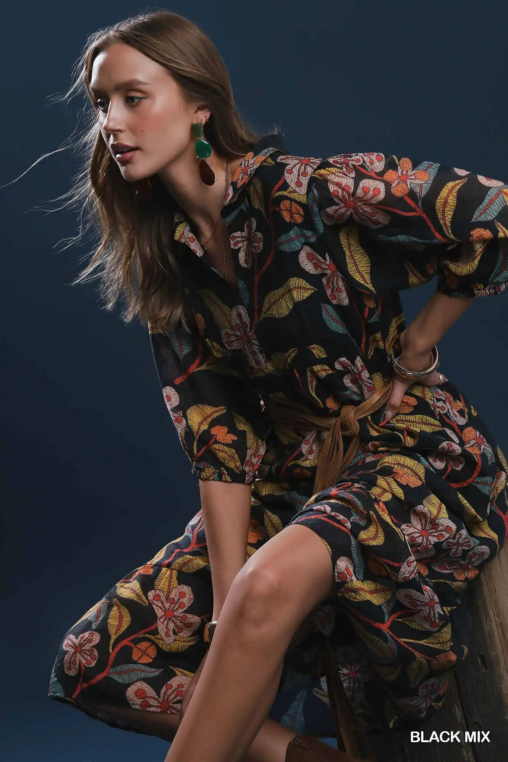 A woman models a black floral print shirt dress with orange, yellow, and green botanical patterns, sitting on a wooden stool against a dark blue background.