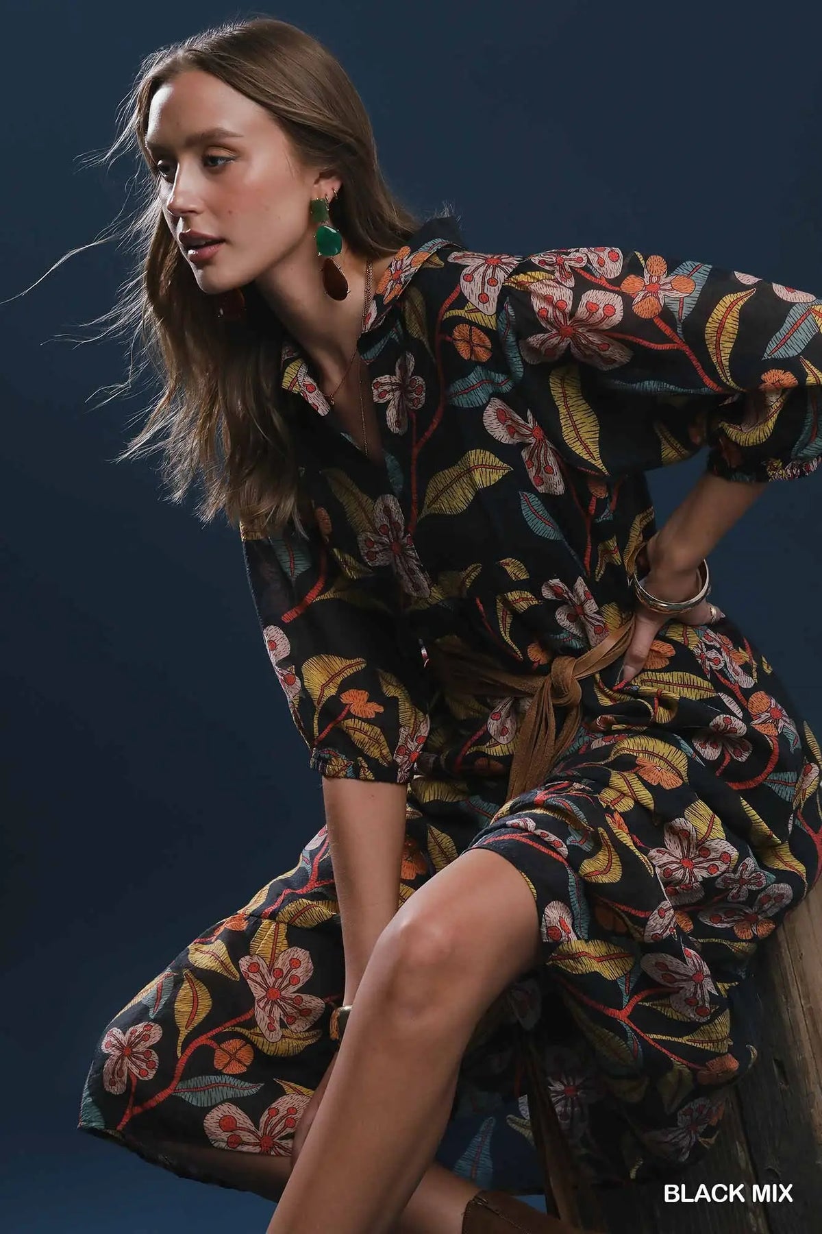 A woman models a black floral print shirt dress with orange, yellow, and green botanical patterns, sitting on a wooden stool against a dark blue background.