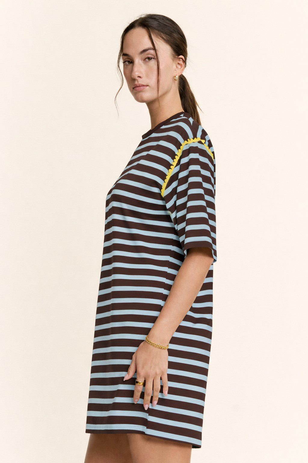 A woman modeling a short-sleeve, crew-neck t-shirt dress with horizontal dark brown and light blue stripes, featuring a yellow braided trim detail along the shoulder, set against a solid cream background.