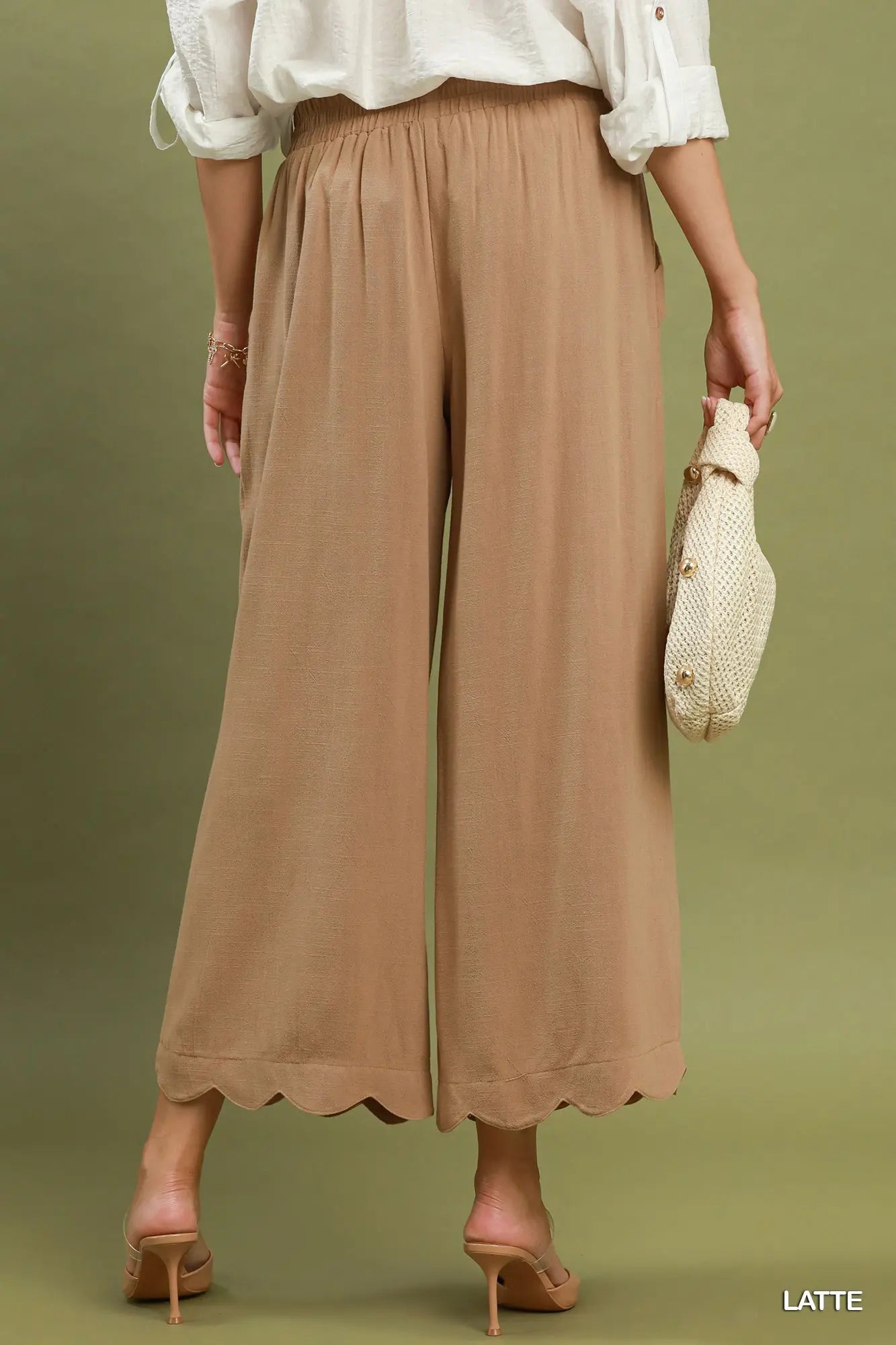 Person wearing brown wide-leg pants and a white blouse, holding a beige handbag on a green background.