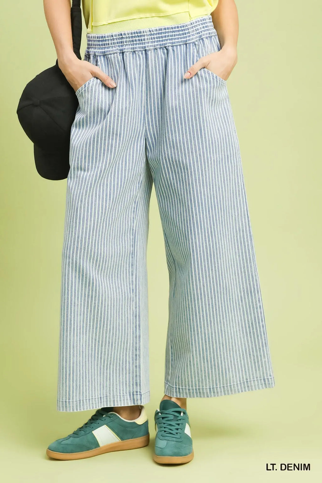 A model wearing light blue and white vertically striped wide-leg cropped pants, styled with teal sneakers and a black cap against a pale yellow-green background.