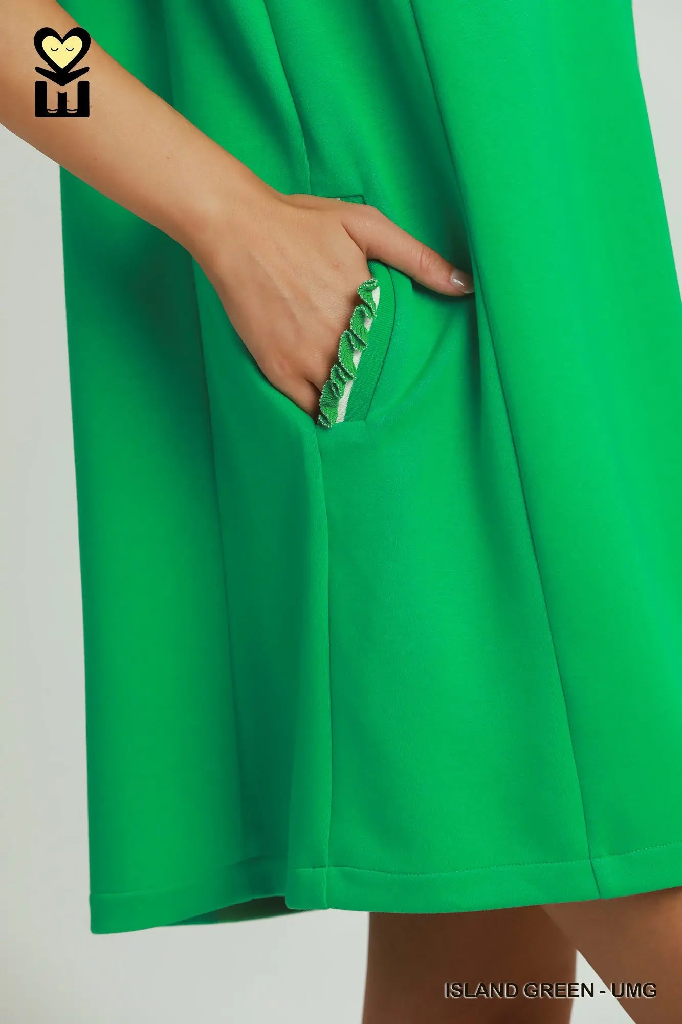 Close-up of a bright green dress with a side pocket, showing a hand reaching into the pocket. The brand logo 'MU' is visible in the top-left corner, and the text 'ISLAND GREEN - UNG' is in the bottom-right corner.