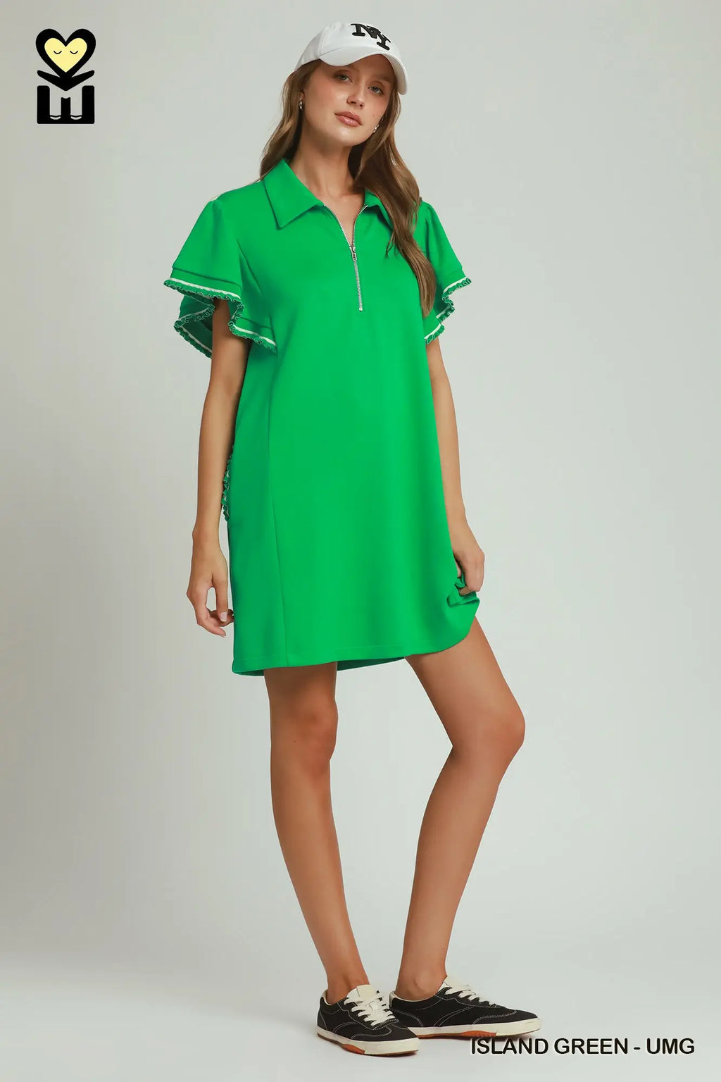 A woman modeling a bright green short-sleeve polo dress with flutter sleeves and a half-zip neckline, paired with a white cap and black sneakers, against a light gray studio background.
