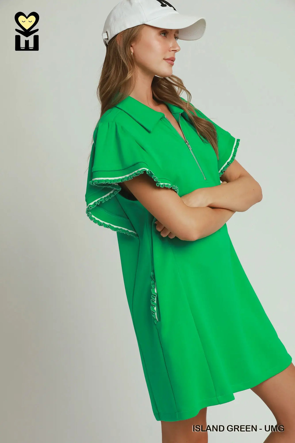 A woman wearing a bright green short-sleeve collared dress with white trim and a white baseball cap, posed against a light gray studio background. The text 'ISLAND GREEN - UMG' appears at the bottom.