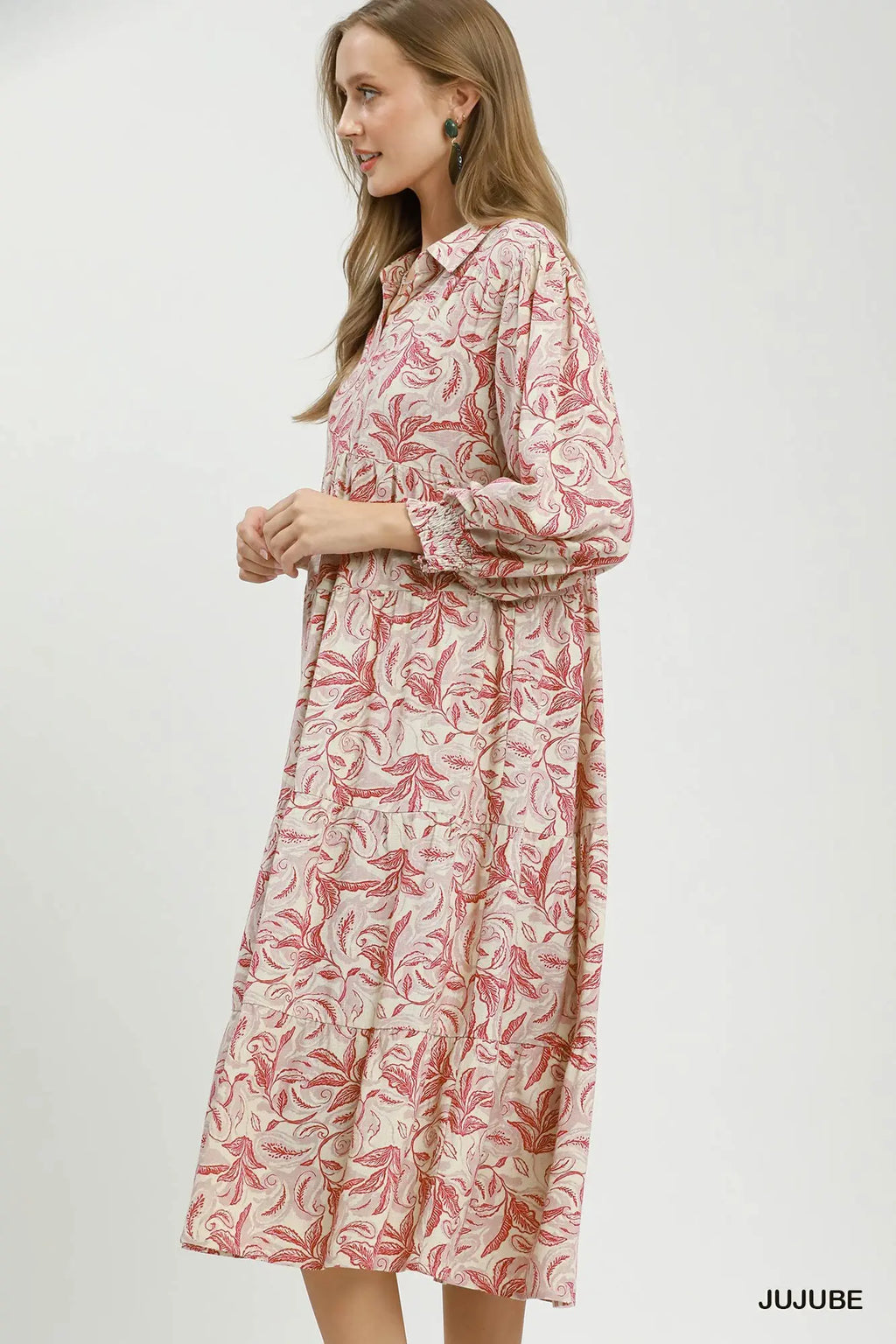 A woman wearing a long-sleeve collared midi dress with a pink and red botanical leaf pattern on a cream background, standing against a plain light gray studio backdrop. The brand name 'JUJUBE' is printed in black in the bottom right corner.