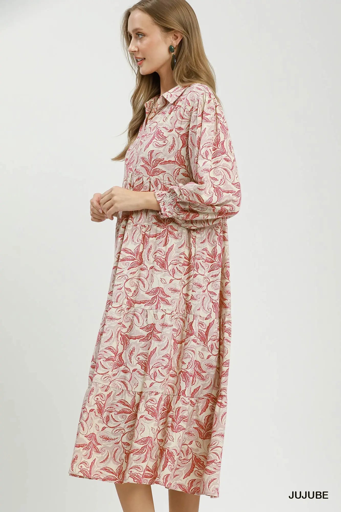 A woman wearing a long-sleeve collared midi dress with a pink and red botanical leaf pattern on a cream background, standing against a plain light gray studio backdrop. The brand name 'JUJUBE' is printed in black in the bottom right corner.