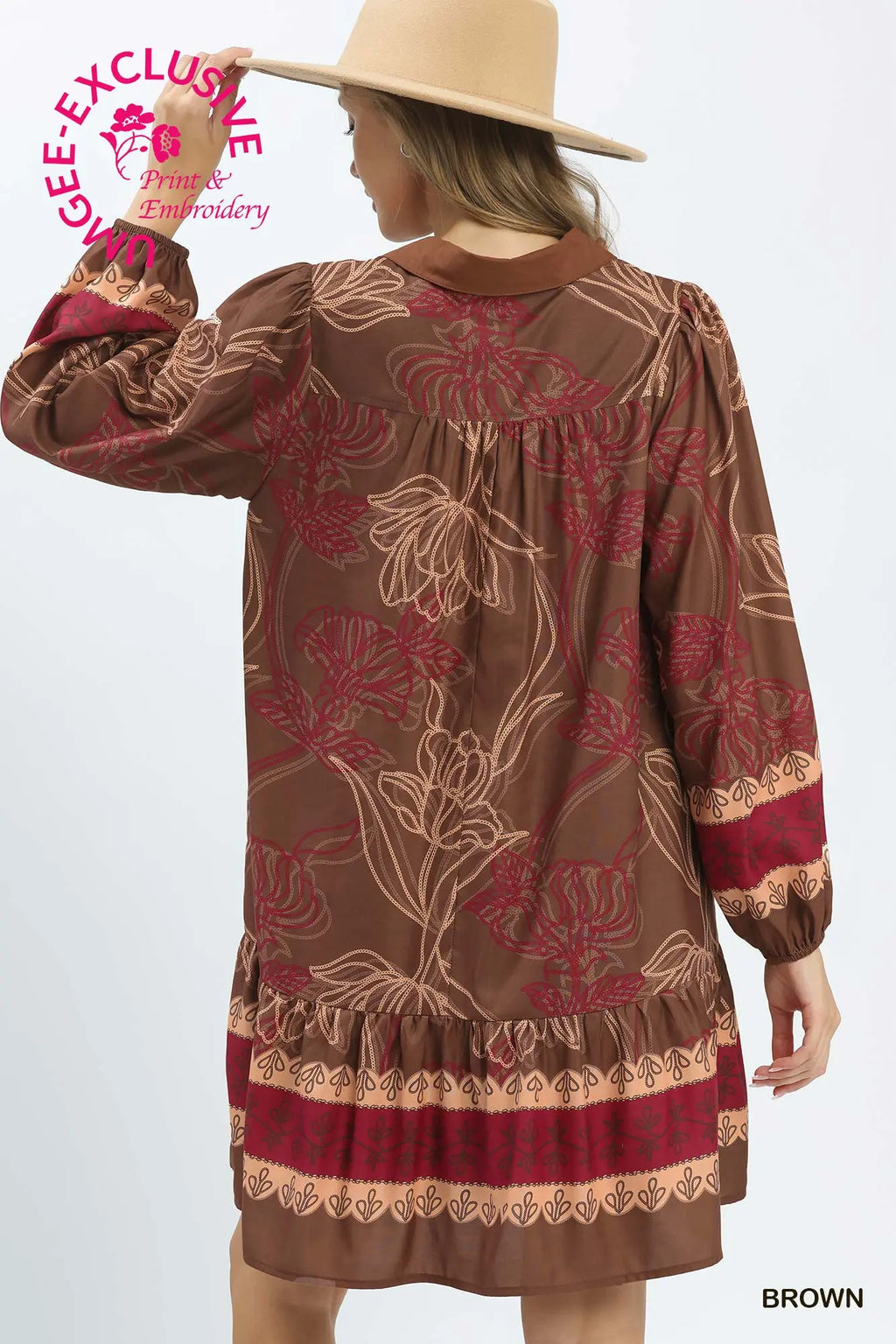 A woman modeling a brown long-sleeve dress with a floral pattern in red and beige, featuring a ruffled hem and cuffs. She is wearing a wide-brimmed beige hat against a plain white background.