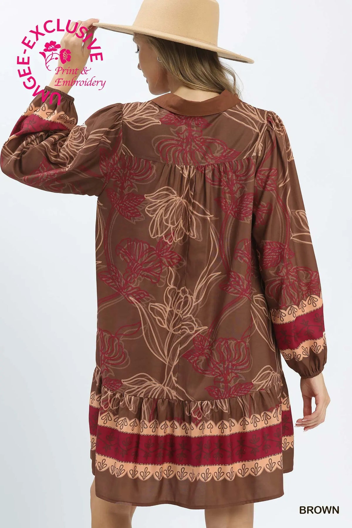 A woman modeling a brown long-sleeve dress with a floral pattern in red and beige, featuring a ruffled hem and cuffs. She is wearing a wide-brimmed beige hat against a plain white background.